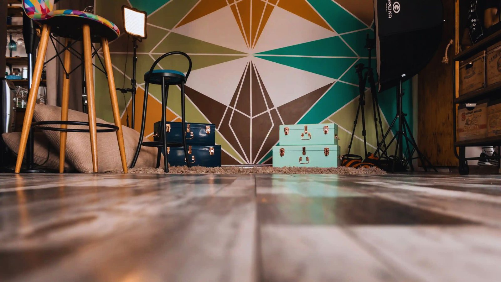 Colorful geometric mural as backdrop with a wooden floor, a stool, and stacked vintage suitcases. Warm, creative studio setting.