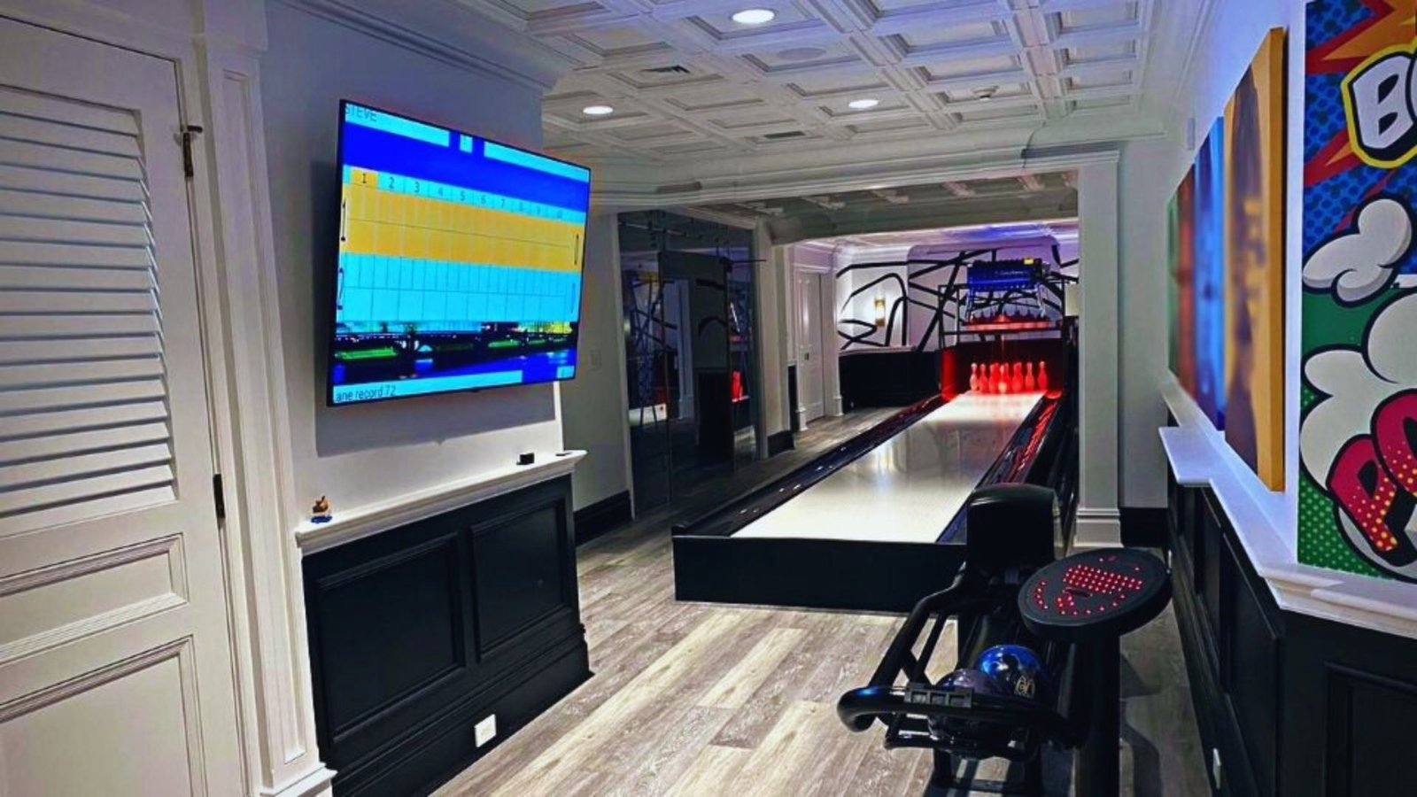 A modern home bowling alley with a sleek wooden floor, brightly lit pins, and vibrant pop art on the walls; a scoreboard displays game scores.