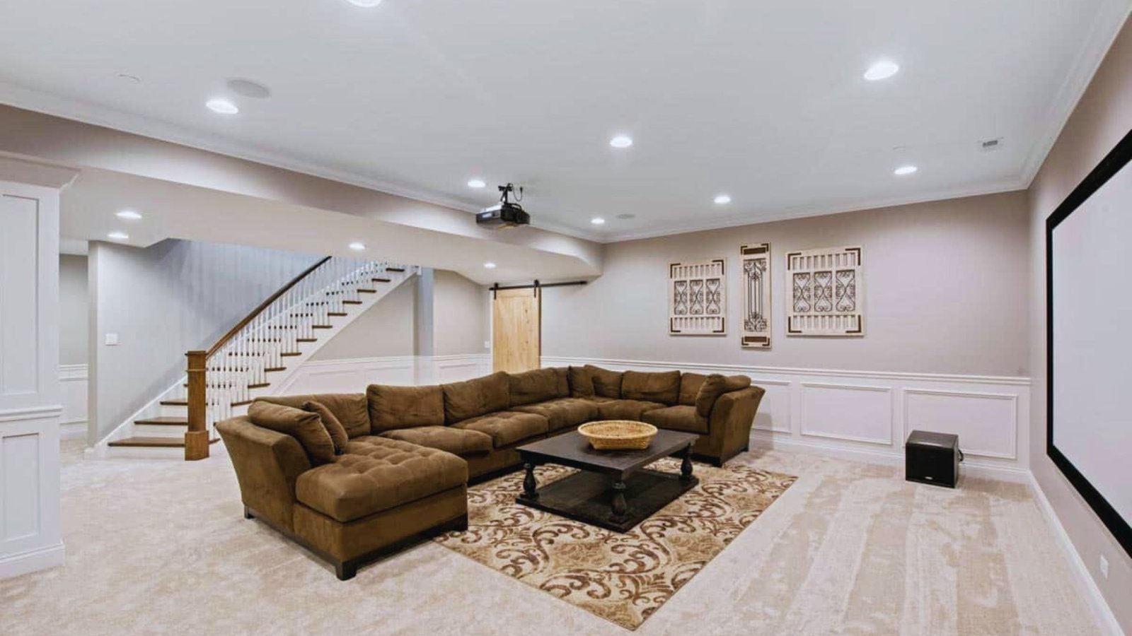 Modern basement entertainment room with a large brown sectional sofa, patterned rug, and a wall-mounted projector screen. Soft lighting adds a cozy atmosphere.