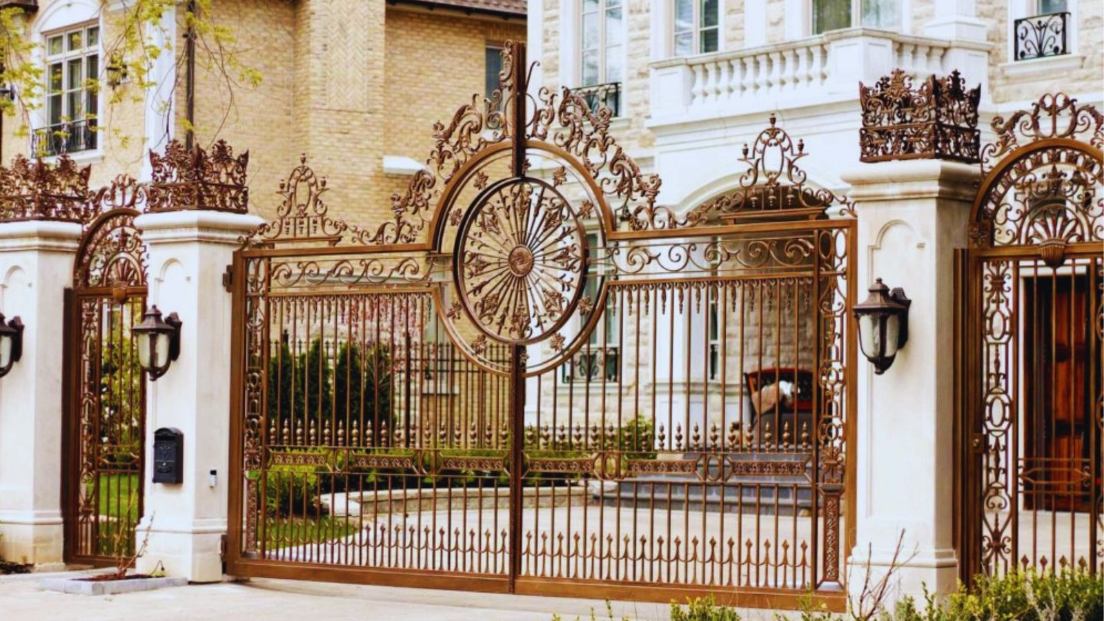 Ornate wrought iron gate with intricate floral patterns and circular design, set between white stone pillars in front of a large, elegant mansion.