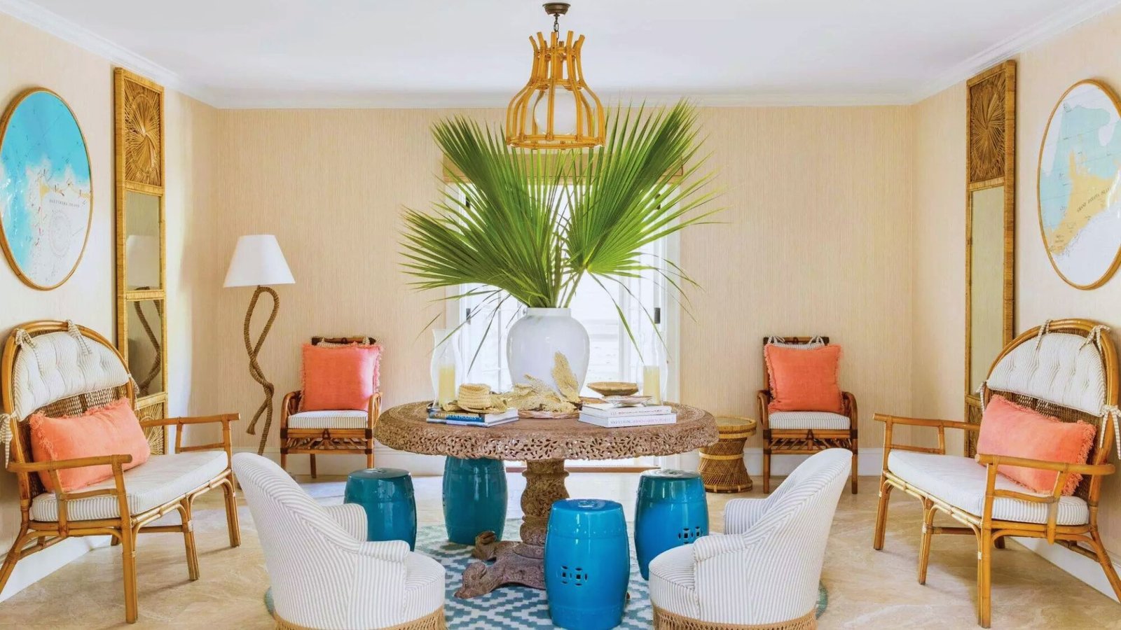 Bright living room with a coastal theme. A round table with a vase of large palm leaves is surrounded by wicker chairs. Blue accents and coral pillows add contrast.