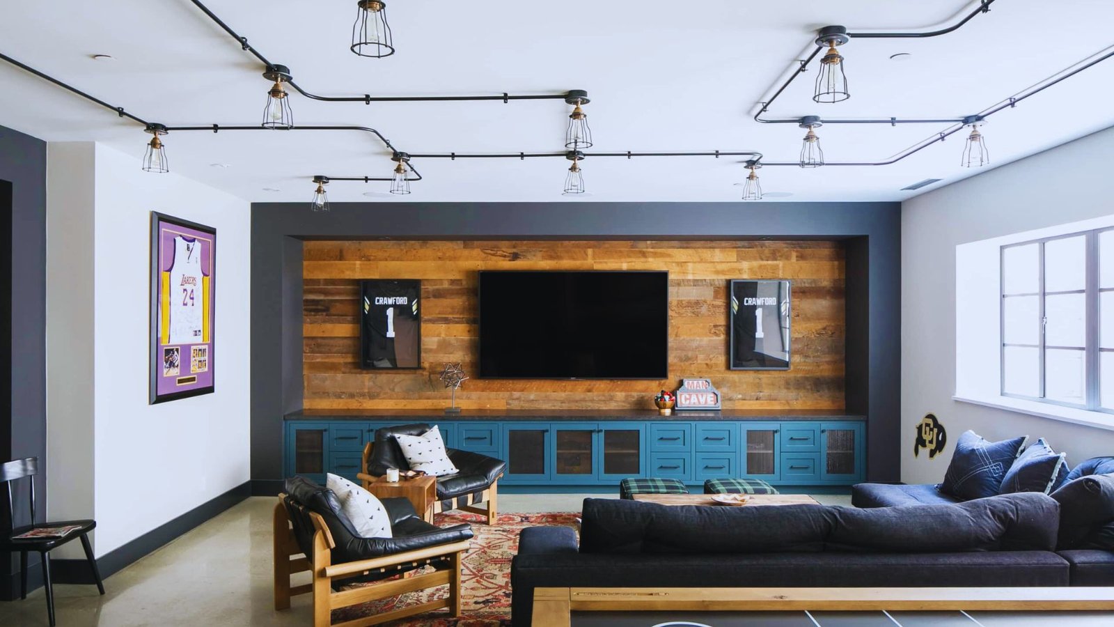 A modern sports-themed living room features a wall-mounted TV on a wooden accent wall, sports jerseys framed on each side, blue cabinets, a dark sectional sofa, and ceiling pipe lights, creating a cozy, sporty atmosphere.