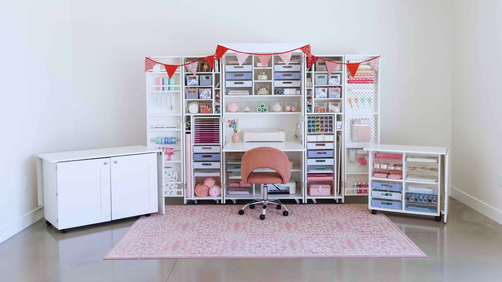 Organized craft room with a pink chair, white shelves full of bins, and colorful crafting supplies. A red bunting hangs above. Bright and inviting.