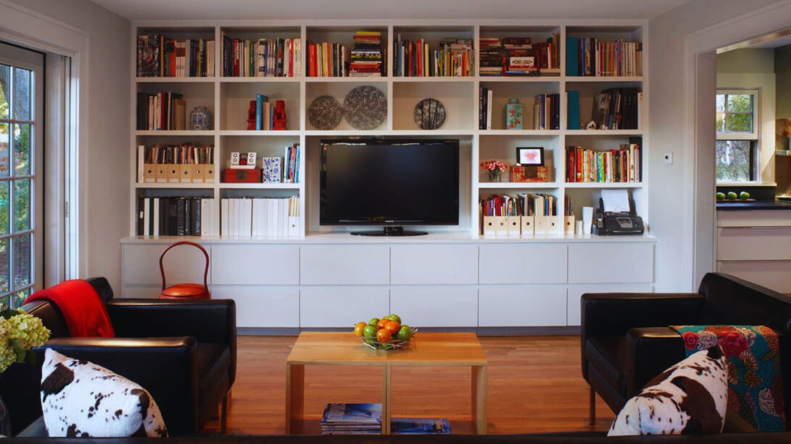 A cozy living room with a white shelving unit filled with books, decor, and a TV. Black chairs have patterned cushions, and a wooden table holds fruit.