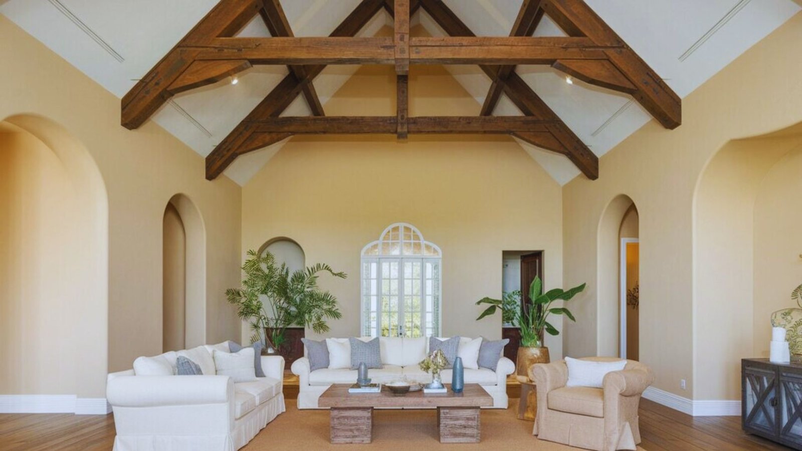 Spacious living room with vaulted ceiling and exposed wooden beams. Neutral tones, white sofas, armchairs, and lush plants create a cozy atmosphere.