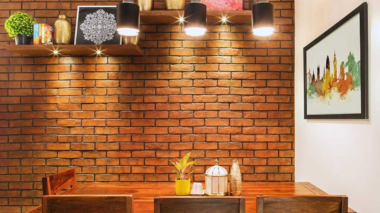 Cozy dining area with a brick wall, wooden table, and two chairs. Shelves with plants and decor are lit by pendant lights, and colorful art hangs nearby.