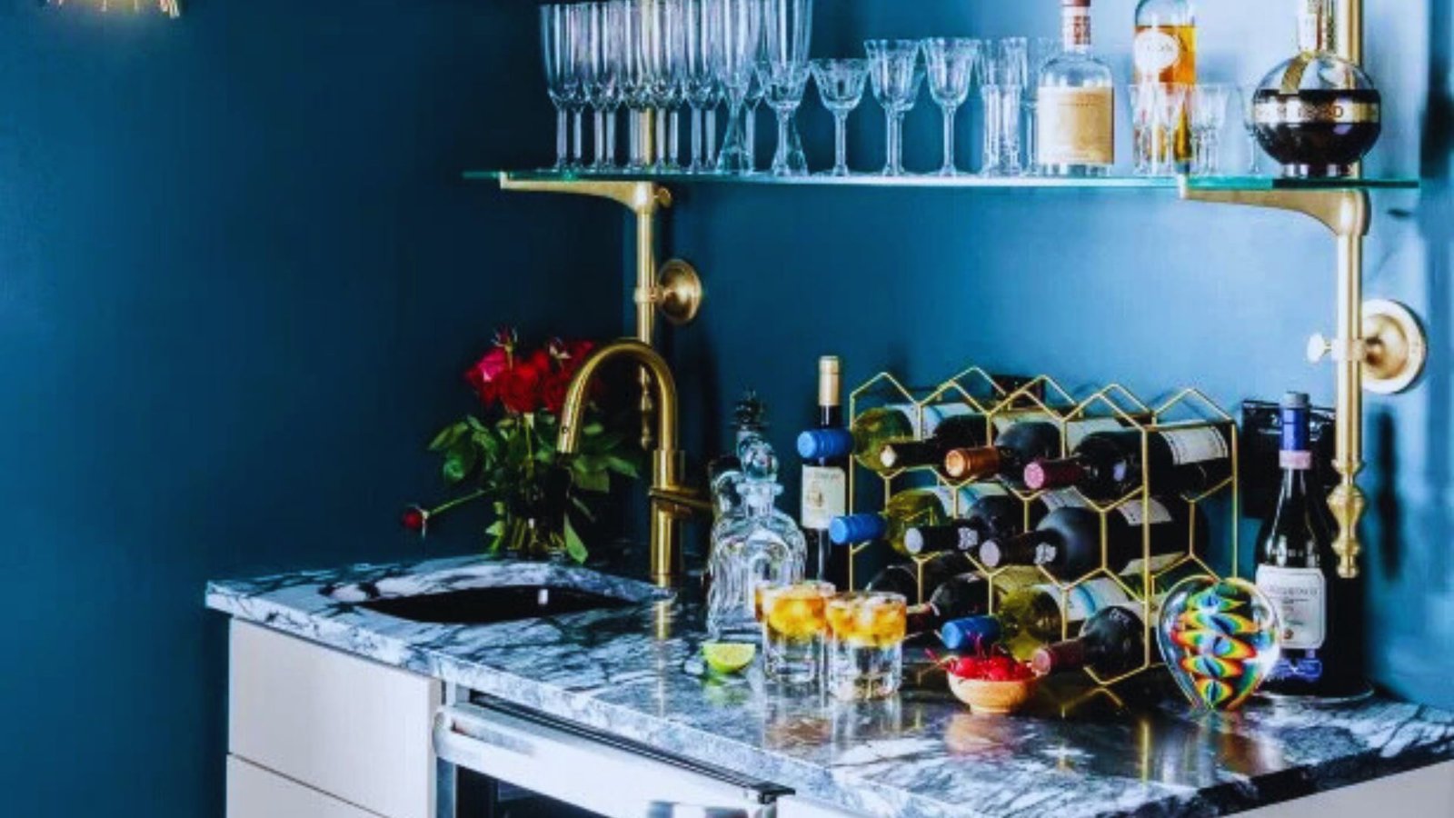 Elegant home bar with a marble countertop, glasses on the shelf, wine bottles in a rack, a golden faucet, red roses, and vibrant bowls, evoking sophistication.