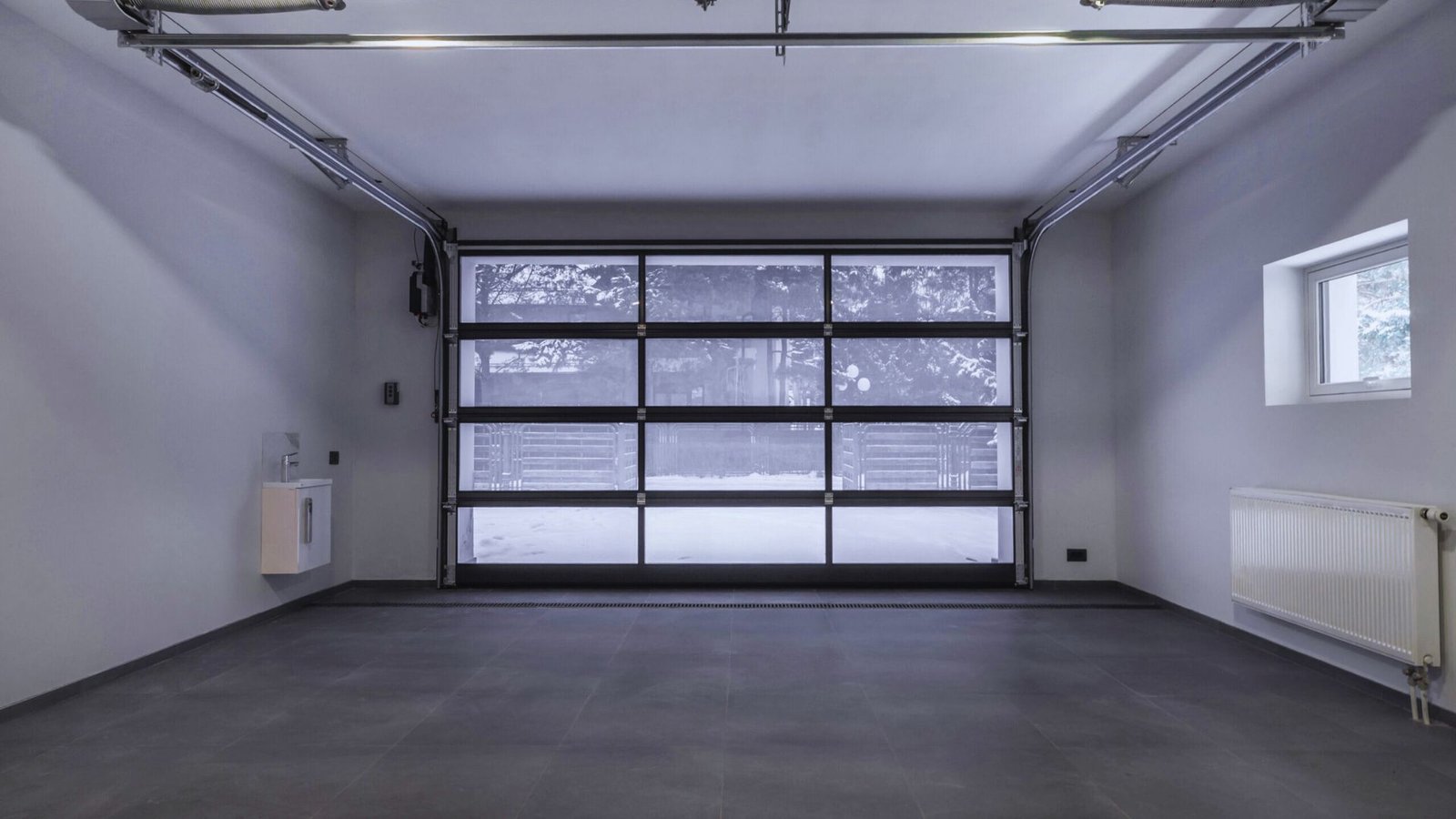 A modern, empty garage with a tiled floor and large glass-panel door. The space is clean and minimalistic, with a window and radiator on the right.