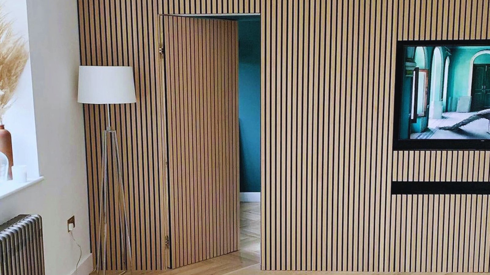 A modern room features a wooden slat wall with a hidden door, revealing a blue interior. A floor lamp stands nearby, and a TV is mounted on the wall.