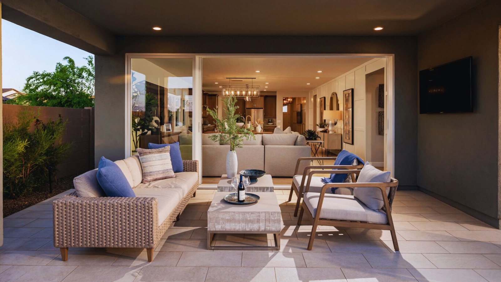 Stylish patio with wicker chairs, blue cushions, and a coffee table. Wide glass doors open to a modern living room, creating a cozy, inviting atmosphere.