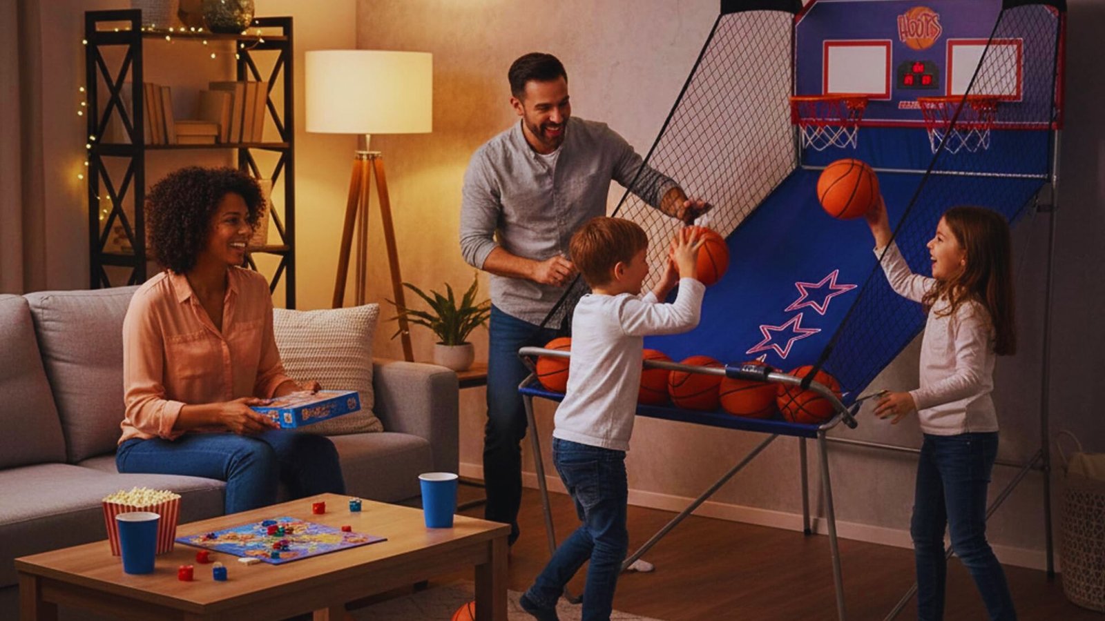 A family enjoys a playful moment indoors: a man and two children play an arcade basketball game while a woman smiles nearby holding a board game.