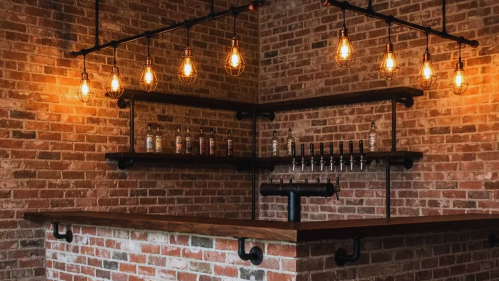 Industrial-style bar with exposed brick walls, warm Edison bulb lighting, and wooden shelves displaying bottles. A row of beer taps is visible. Cozy atmosphere.