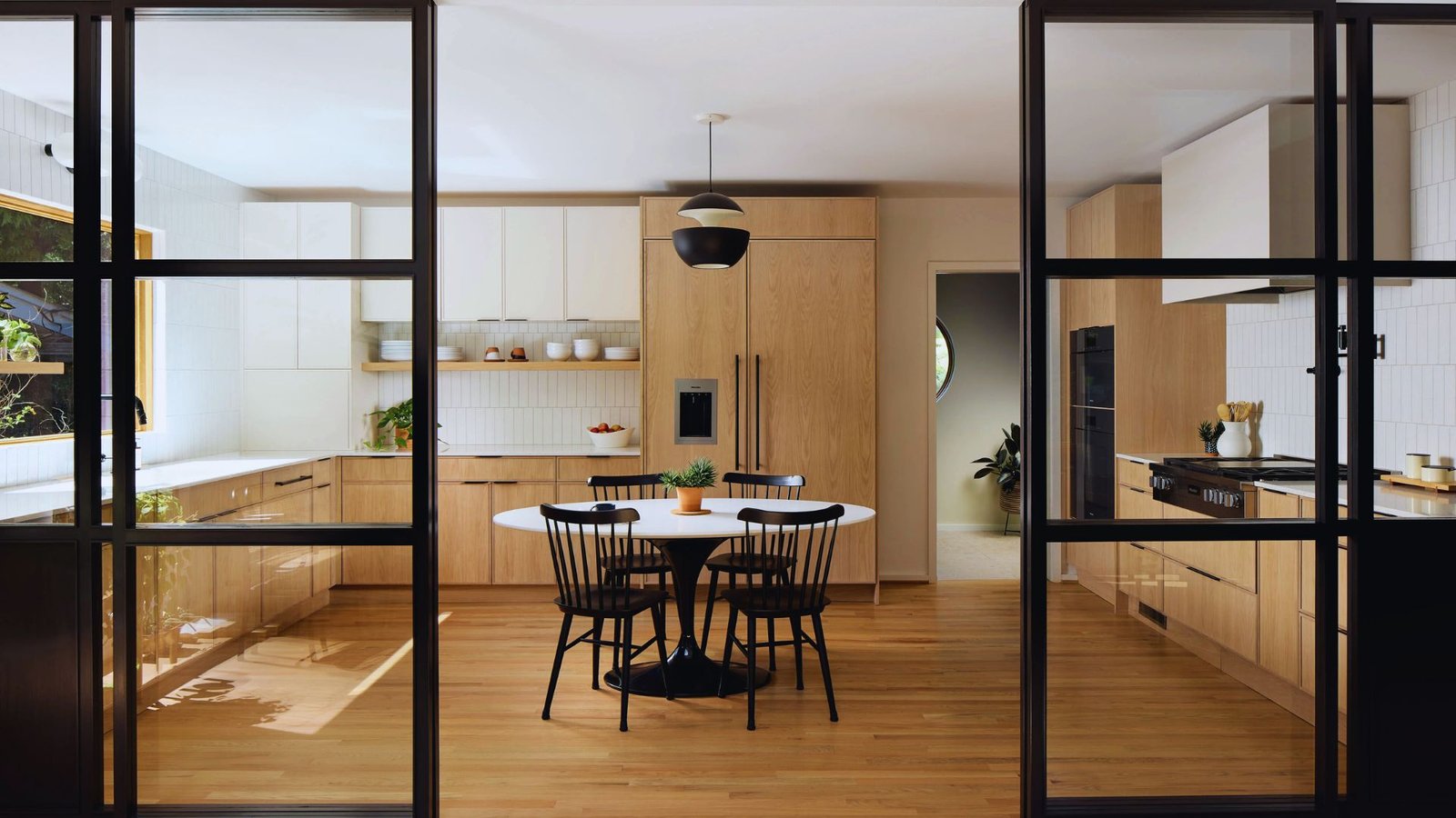 Modern kitchen with wooden cabinets, a round white table, and black chairs. Natural light streams in from a window, creating a warm, inviting atmosphere.