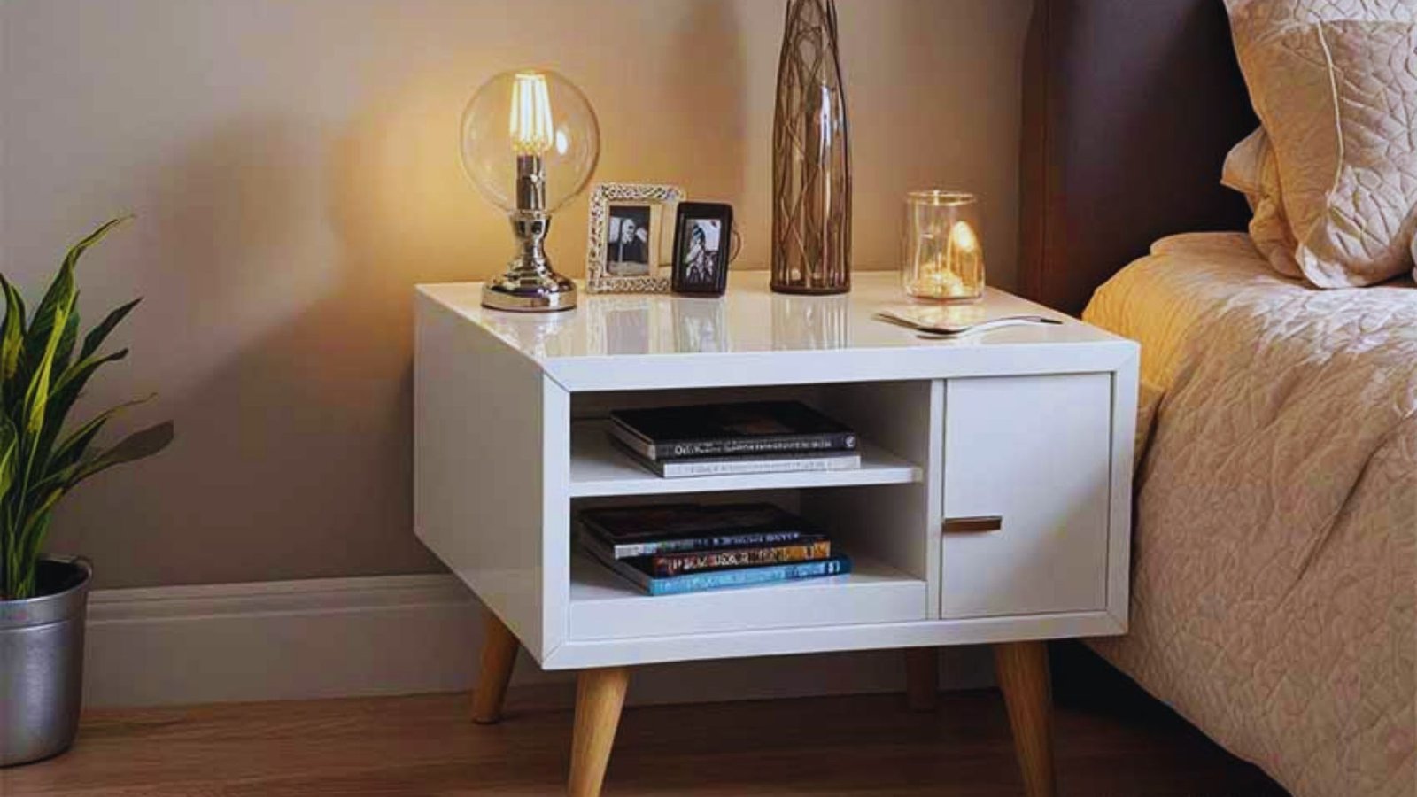 A cozy bedside scene with a modern white nightstand featuring a glowing lamp, books on shelves, framed photos, and a plant, creating a warm, inviting tone.