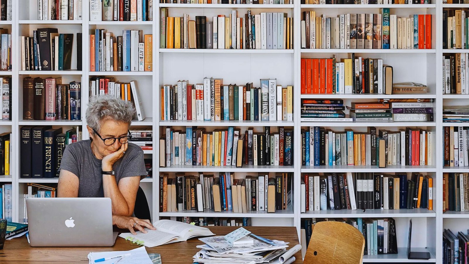  A person reads at a wooden table with a laptop and papers, surrounded by a large, colorful bookshelf filled with assorted books, conveying focus and concentration.