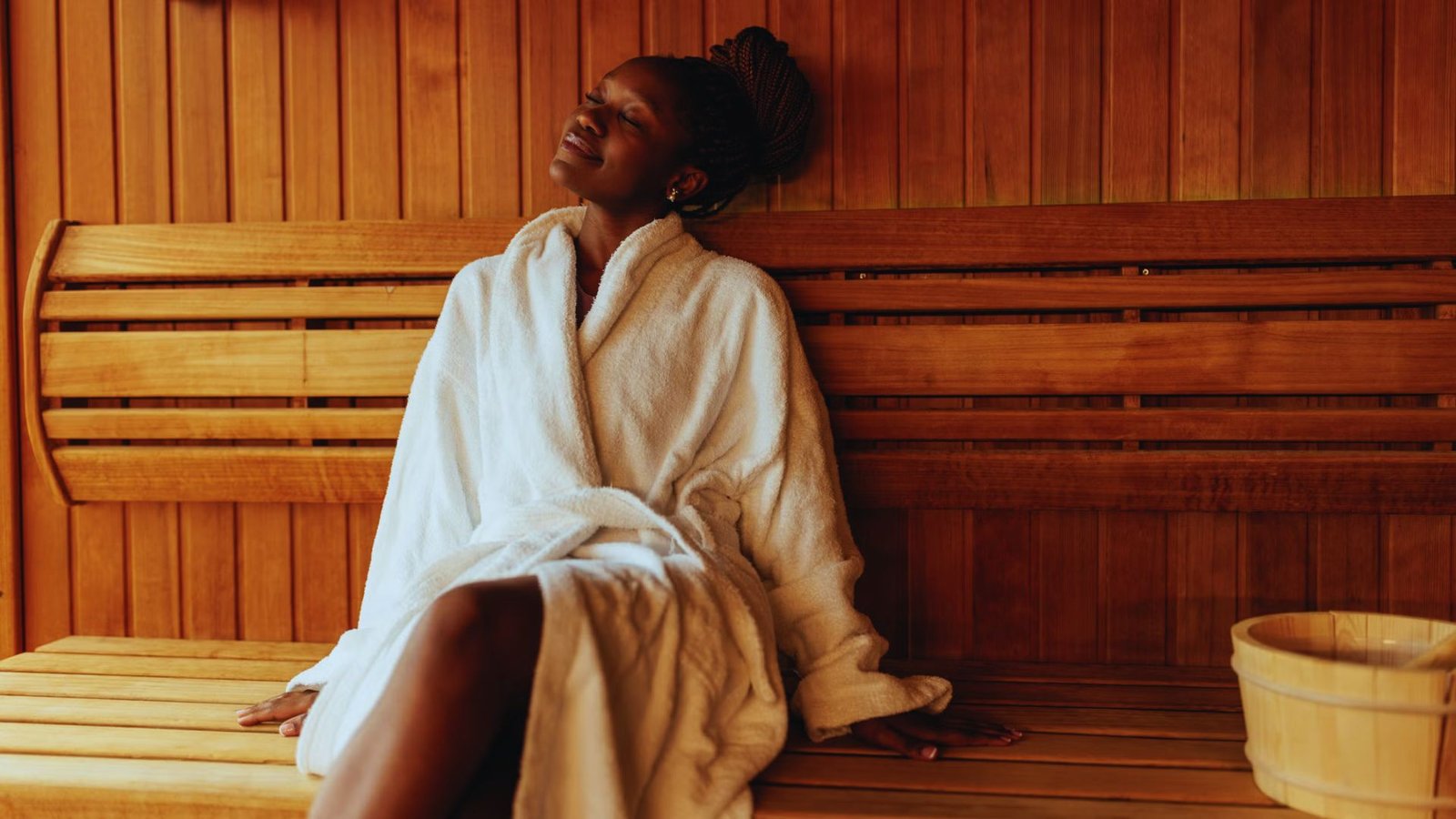 A woman in a white robe relaxes in a wooden sauna, eyes closed, with a peaceful expression. A bucket is placed beside her on the bench.