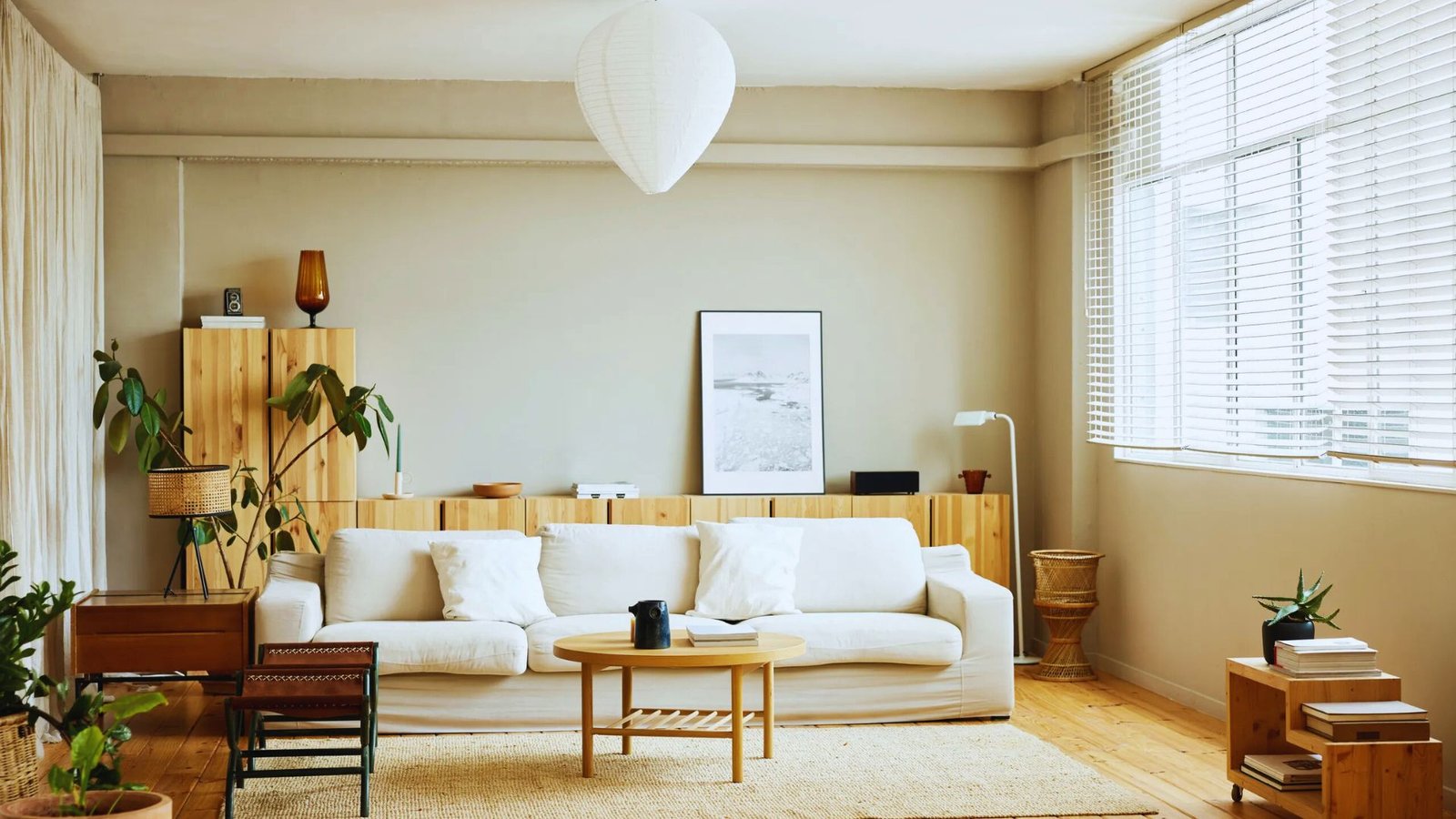 Cozy living room with a white sofa, wooden coffee table, and potted plants. Bright natural light from large windows enhances the warm, minimalistic decor.