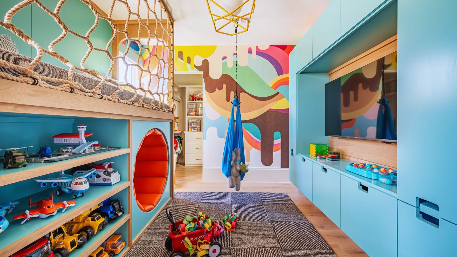 Colorful children's playroom with a loft bed, rope netting, and vibrant mural. Shelves hold toy vehicles. A blue swing hangs in the center, evoking joy.