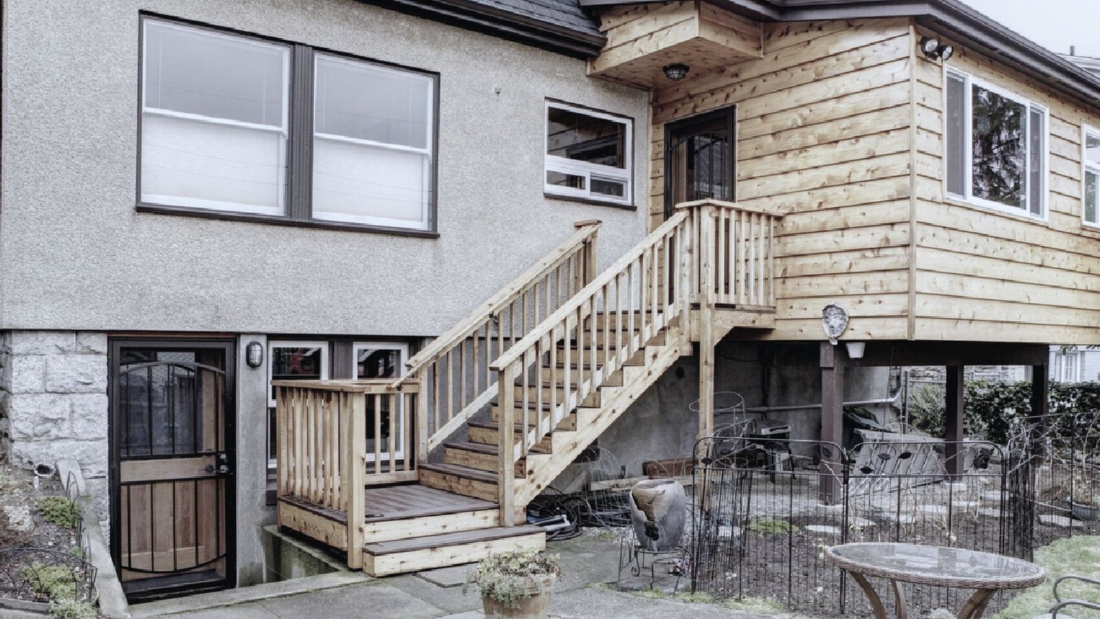 The image shows a two-story house exterior with a gray lower half and wooden upper extension. A wooden staircase leads to the second floor, with a small fenced garden area below. The overall tone is neutral, with natural wood textures and a simple, rustic atmosphere.
