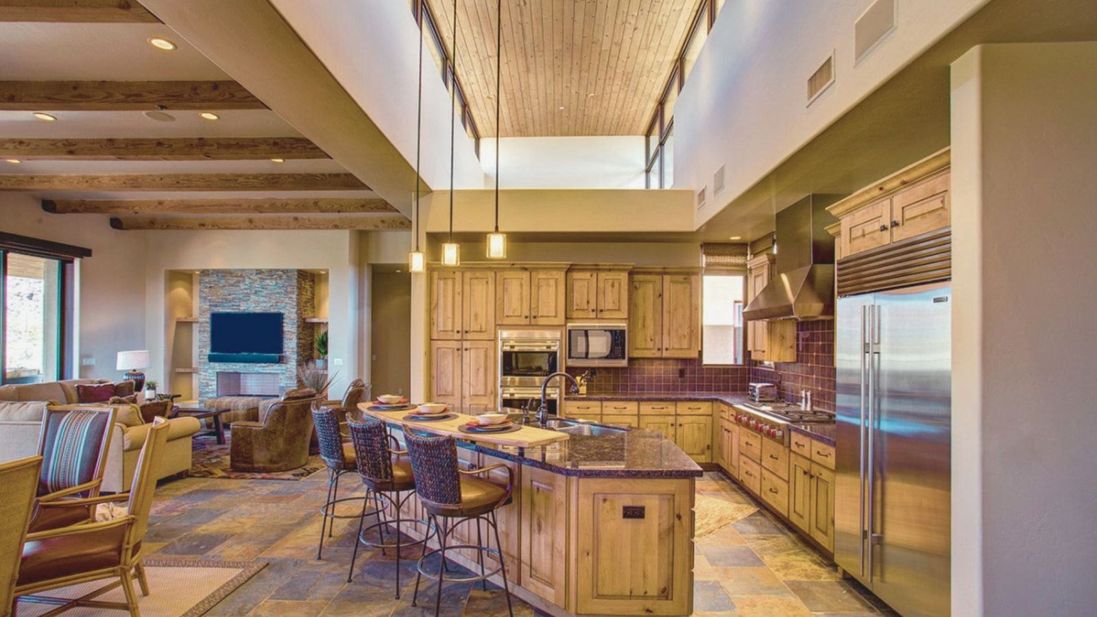 Spacious open-concept kitchen and living room with warm lighting. Features wooden cabinets, granite countertops, stone flooring, and cozy seating.