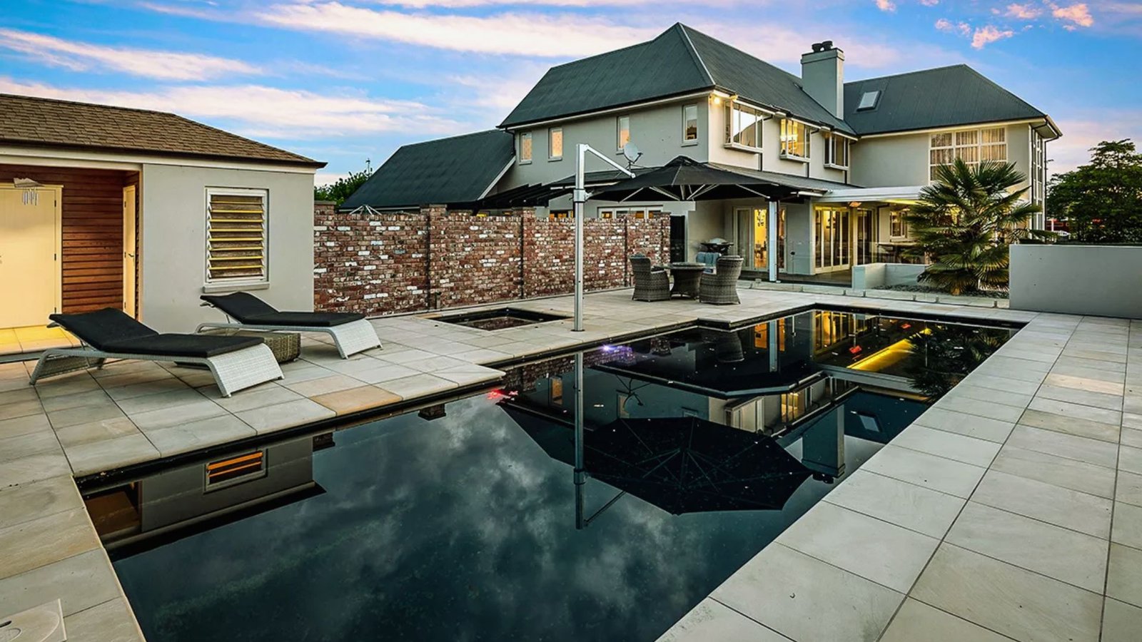 A house featuring a pool and a spacious patio area, surrounded by greenery and outdoor furniture.
