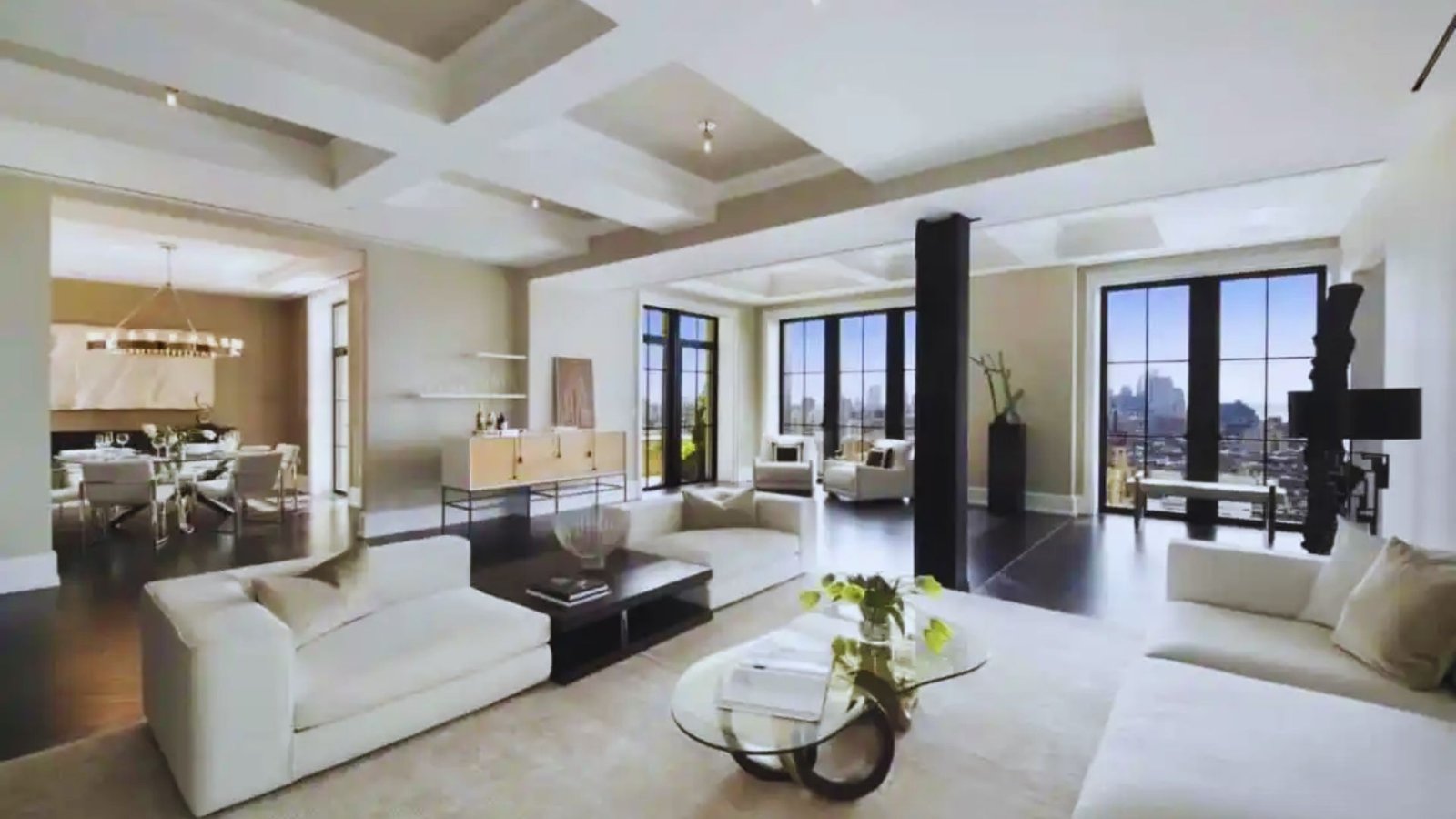 A bright living room featuring white furniture and large windows, allowing natural light to fill the space.